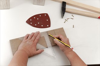 A woman's hands marking lines on a piece of wood with a square and a pencil on a white table in her home woodworking shop