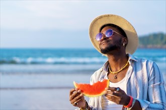 Black ethnic man enjoy summer vacation at the beach eating a watermelon with a sunshade and sunglasses
