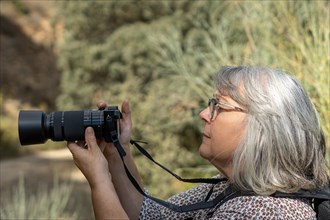 Older woman photographer with white hair and glasses taking a picture looking through the viewfinder of her camera