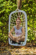 Smiling entrepreneur at her juice shop backyard resting on swing chair while drinking fresh detox juice