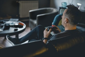 An adult man sitting on a sofa and holding a joystick while playing a video game on console