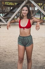 Beautiful young green eyed woman in red bikini and shorts looking at camera standing on beach. Mysterious and attractive look. Vertical portrait
