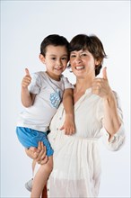 Young mother and son standing together on white background making happy thumbs up hand gesture. approving expression looking at camera showing success