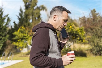 Adult man listening to messages on his phone and drinking mate infusion while walking in his garden. Home office concept