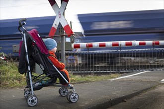 Subject: Traffic. Child in a pram in front of a level crossing with a goods train behind