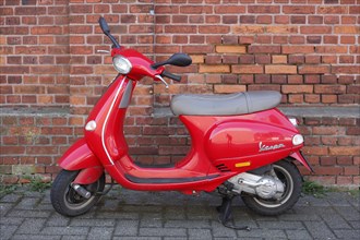 Red Vespa scooter standing in front of a reddish brick wall on the footpath
