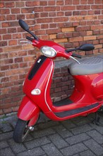 Red Vespa scooter standing in front of a reddish brick wall on the footpath