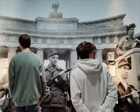 Two young people from today stand in front of a photo of the construction of the Wall with young soldiers