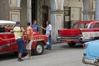 Cuban men discussing on street and red old 1950s vintage American cars