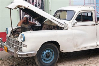 Cuban car mechanic repairing 1950s vintage American Chrysler pickup truck on street in Trinidad