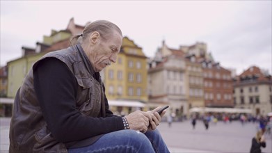 Senior sits on the steps and uses a smartphone in the historic center of an old European city