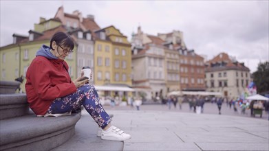Elderly woman sits on the steps drinking coffee and using a smartphone in the historic center of an old European city