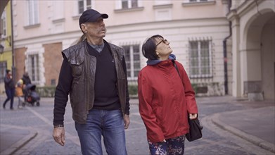 Elderly couple of tourists are walking through the historical center seeing the sights and looks at up in an old European city