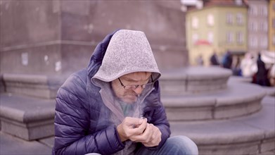 Adult man ? ????? sitting on square and lights up at a tobacco pipe releasing smoke in the Palace Square
