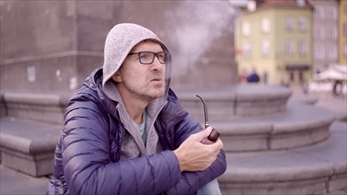 Adult man with glasses sitting on square and smoking a tobacco pipe releasing smoke in the Palace Square