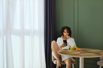 Young woman in bathrobe using smartphone and eats grapes after taking bath