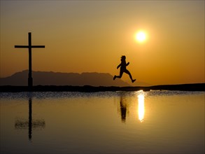 Silhouette of a woman jumping for joy next to a summit cross at sunset