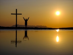 Silhouette of a woman with arms stretched up next to a cross on a summit at sunset