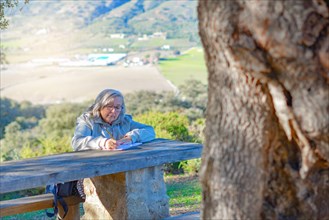 Woman sitting on a bench writing in a notebook with a sunlit mountain scenery in the background