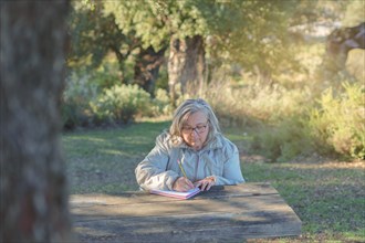 Front view of a white-haired woman sitting at a wooden table in a park writing in a notebook