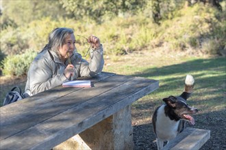 White-haired woman sitting at a park table with her glasses in her hands writing in a notebook accompanied by her dog
