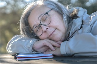 Woman with white hair and glasses sleeping on a notebook on a wooden table dreaming with happy face
