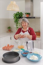 Older white-haired woman in apron cracking eggs to make a cake in her kitchen