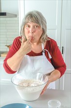 Older white-haired woman cook in apron sucking her finger tasting the cream on her strawberry cake