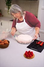 White-haired female pastry chef in an apron decorating a strawberry cake in her kitchen