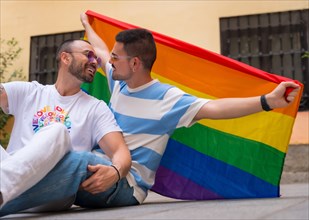 Portrait of gay male couple sitting on the floor with rainbow flag at pride party