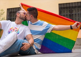 Portrait of gay male couple sitting on the floor kissing with rainbow flag at pride party