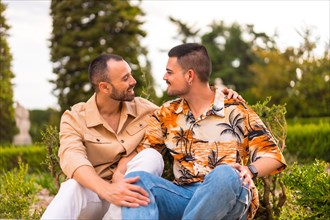 Romantic portrait of gay newlyweds sitting kissing at sunset in a park in the city