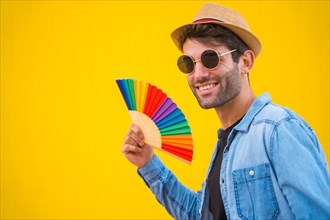 Portrait of a caucasian man in a hat smiling with a rainbow lgbt fan on a yellow background