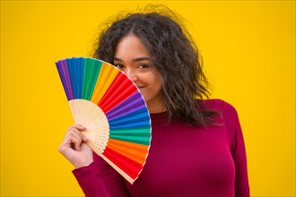 Portrait of a latin woman smiling with a rainbow lgbt fan on a yellow background