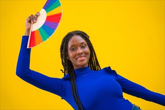 Portrait of a black ethnic woman dancing with a rainbow lgbt fan on a yellow background