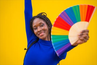 Portrait of an attractive black ethnic woman with a rainbow lgbt fan on a yellow background