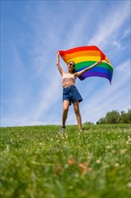 Caucasian brunette woman with a rainbow lgbt flag on the grass and blue sky background