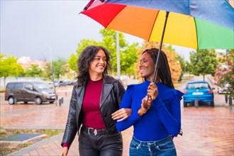 Multiethnic female friends laughing with an umbrella in the rain in a city