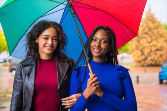 Portrait of laughing multi-ethnic female friends with a rainbow umbrella in the rain in a city