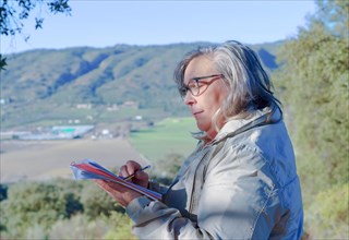 Woman with white hair and glasses writing in a notebook with a pencil in the background of a mountainous landscape