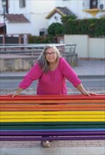 Older woman with white hair and pink shirt standing behind a bench painted with the colors of the LGTBIQ flag smiling at the camera