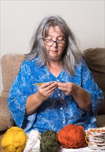 Front view of an older woman with white hair and glasses crocheting with her wool in the foreground
