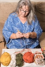 Front view of an older woman with white hair and glasses crocheting with her wool in the foreground
