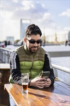 A smiling man sending messages while smiling at a bar near the river