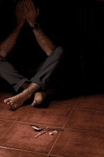 Unidentified barefoot man sitting on the floor in semi-darkness with a syringe and spoon containing heroin in the foreground