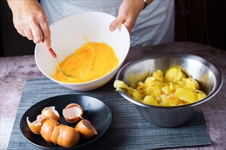 Woman in white apron beating an egg in a bowl with a plate of eggshells and a bowl of potatoes to make an omelet