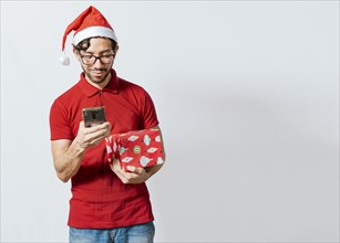 Christmas man holding gift box and telephone isolated. Handsome man in christmas hat holding gift and looking at phone