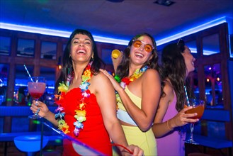 Female friends in a nightclub dancing with glasses of alcohol smiling at a night party on summer vacation