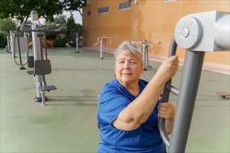 Active obese elderly woman in sportswear working out on a gym machine in a park gym while listening to music on headphones