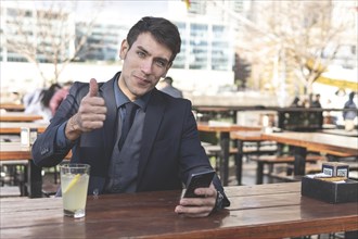 Businessman sitting at an outdoor bar drinking lemonade looking to the camera giving a thumb up